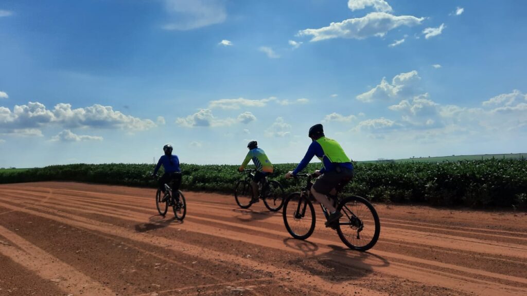 Três pessoas andando de bicicleta durante a atividade promovida pelo Sesc Catanduva. O cenário é uma trilha de terra, com vegetações em um campo aberto ao fundo e céu ensolarado com algumas nuvens. Os ciclistas vestem roupas apropriadas para a prática e capacetes de ciclismo.